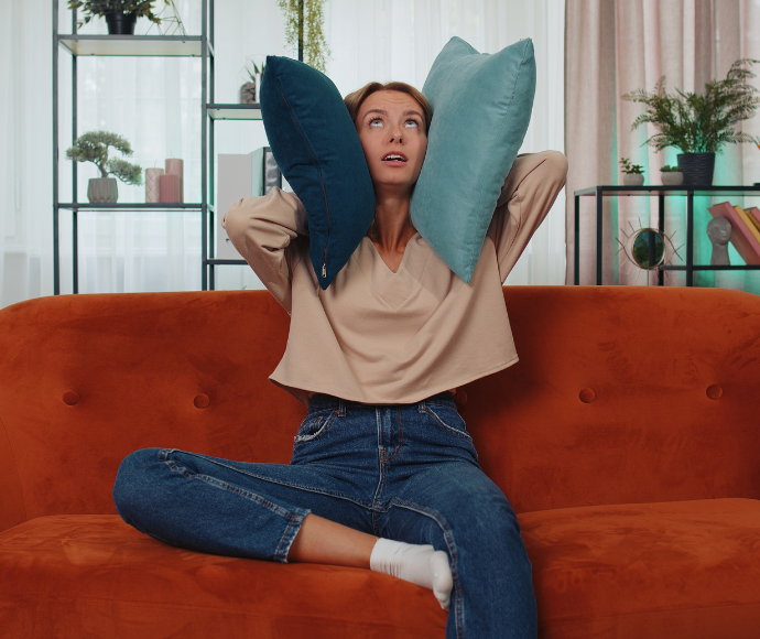 Woman sitting on orange couch with two pillows covering her ears.