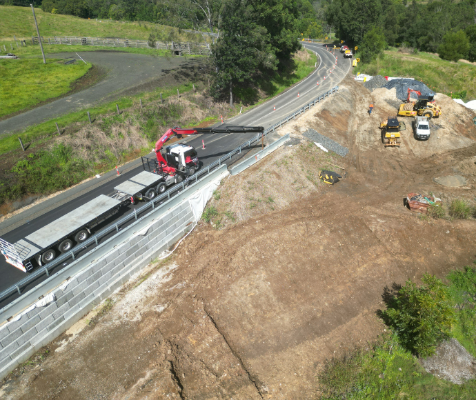 A construction site with heavy machinery is at work alongside a rural road. A red crane is parked on the road, and various construction vehicles are visible on the dirt areas.