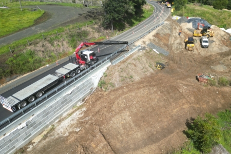 A construction site with heavy machinery is at work alongside a rural road. A red crane is parked on the road, and various construction vehicles are visible on the dirt areas.
