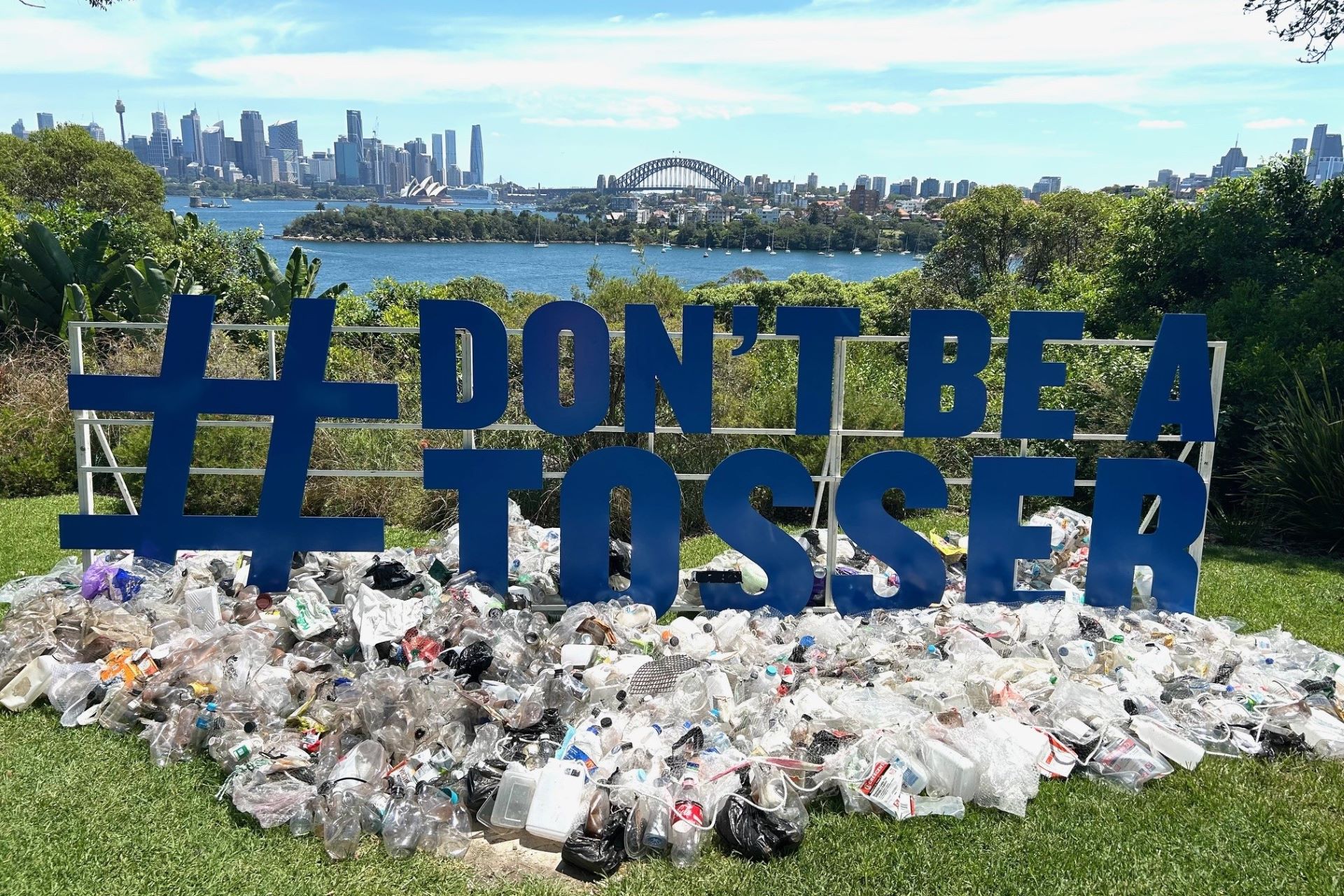 Large Don't Be A Tosser sign with litter spread underneath and Sydney Harbour and the Harbour Bridge in the background