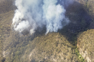 View from above of a bushfire. White smoke billows up towards the camera casting a shadow to the right over brown-green bush, which surrounds the fire on all sides.